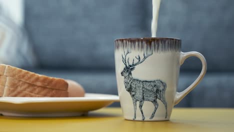 Heiße-Milch-In-Die-Keramiktasse-Mit-Rentiermotiv-Gießen,-Neben-Einem-Teller-Mit-Geschnittenem-Brot