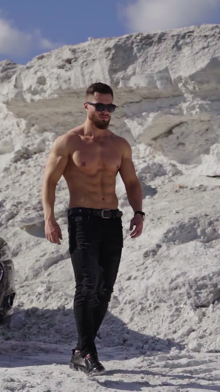 Handsome man near the black car. Shirtless guy in black sunglasses with sportive body walking in white mountains in a summer day. Slow motion. Vertical video