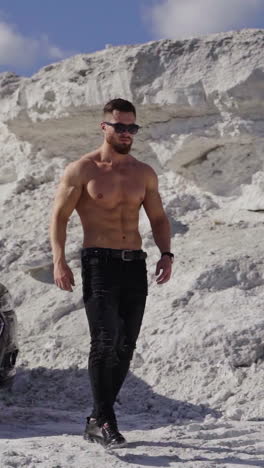 handsome man near the black car. shirtless guy in black sunglasses with sportive body walking in white mountains in a summer day. slow motion. vertical video