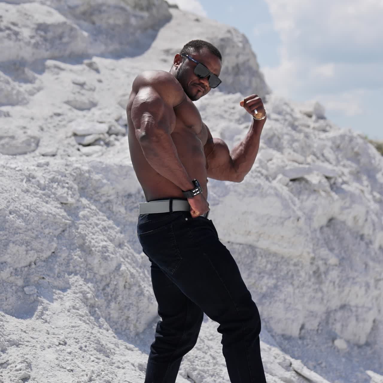 Atleta muscoloso su sfondo roccioso bianco. Uomo afroamericano con occhiali neri senza maglietta che mostra i bicipiti. Vista di profilo
