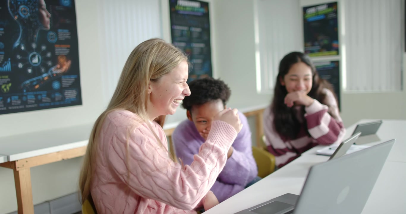 Diverse teenage females reacting to phone content, laughing and leaning at school desk with laptop
