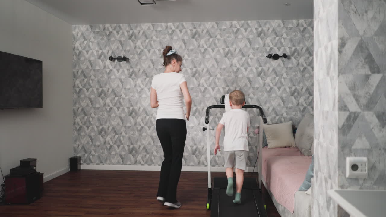 Woman motivates young boy to boost his pace while using treadmill in modern living room with patterned walls and wooden floor, promoting active lifestyle and positive fitness habits in child