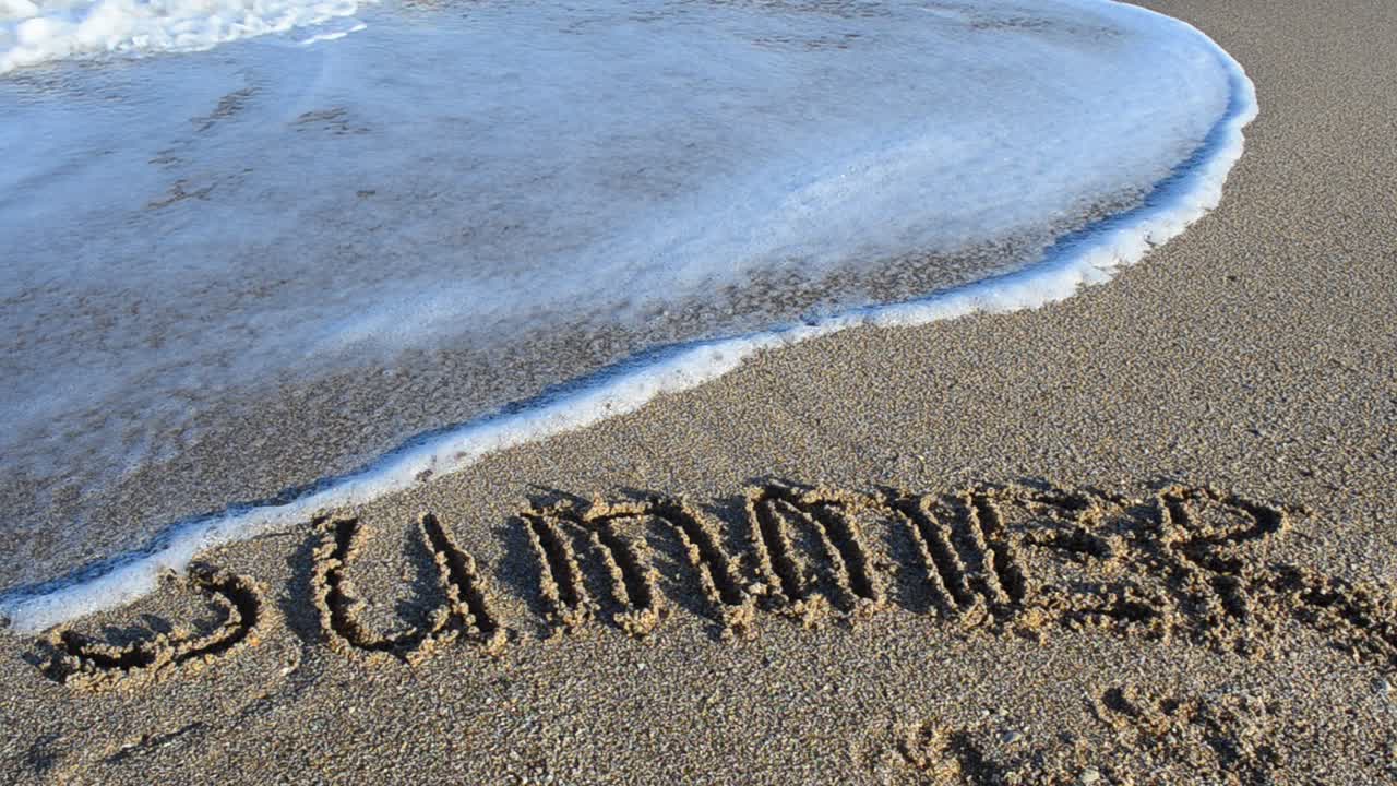 Scritta "estate" sulla sabbia. L'iscrizione viene cancellata da un'onda marina. Riprese in spiaggia.