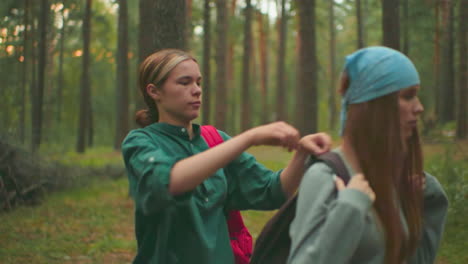 young woman in green shirt assists her friend wearing blue bandana to put on backpack in forest, sunlight filters warmly through trees, illuminating scene as they look at each other in silent