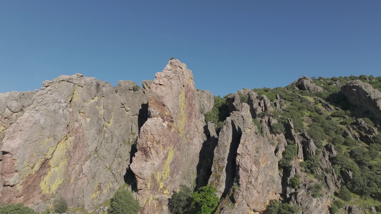Slow-motion, stationary drone shot that raises the camera over multi-colored rock formations with sculpted peaks and a vibrant blue sky in the background, revealing dramatic natural shapes.