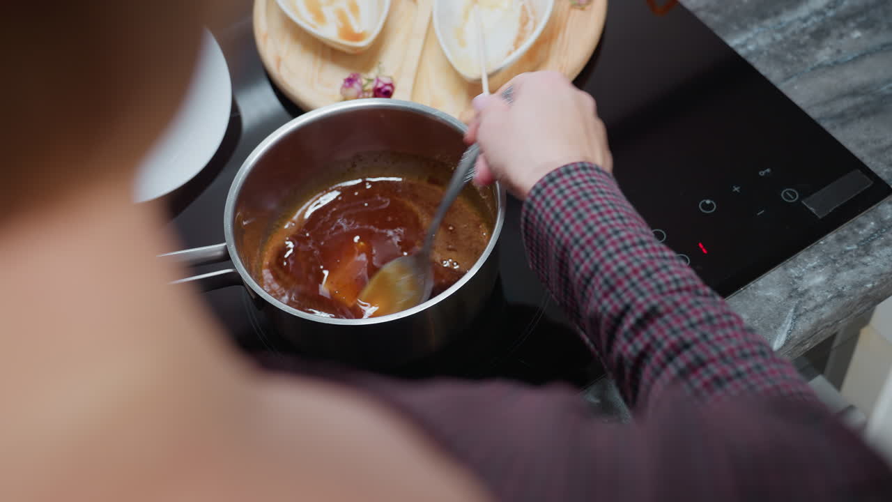 Rear view of person stirring thick soup in stainless steel pot on induction stove while adjusting cooking temperature, with wooden tray, used bowls, and utensils arranged on dark marble countertop