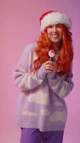 vertical video studio portrait shot of young gen z woman wearing christmas santa hat holding candy lollipop and dancing