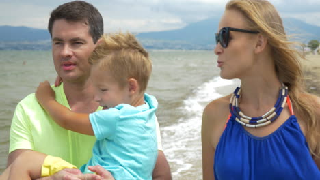 young family is walking along the beach having a lovely conversation and smiling dad carries the baby mother is going near them