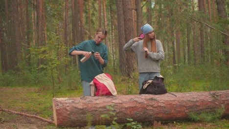 hikers pause in serene forest setting, one fills thermos lid with water, while other brushes hair, both engaged in personal routines, backpacks resting on fallen tree