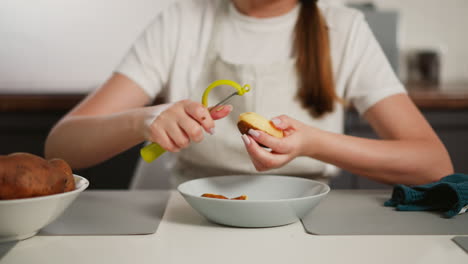 close up of chef peeling raw potato with yellow peeler into bowl on kitchen table, partial view of apron and cloth nearby, clean home kitchen setup with fresh ingredients and focused preparation