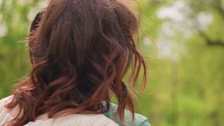 Primo piano di una madre bianca che abbraccia la badante in un parco verde, abbraccio intimo e tocco confortante, movimento delicato dei capelli, gesto di supporto, sfondo di fogliame primaverile, scambio emotivo tra adulti