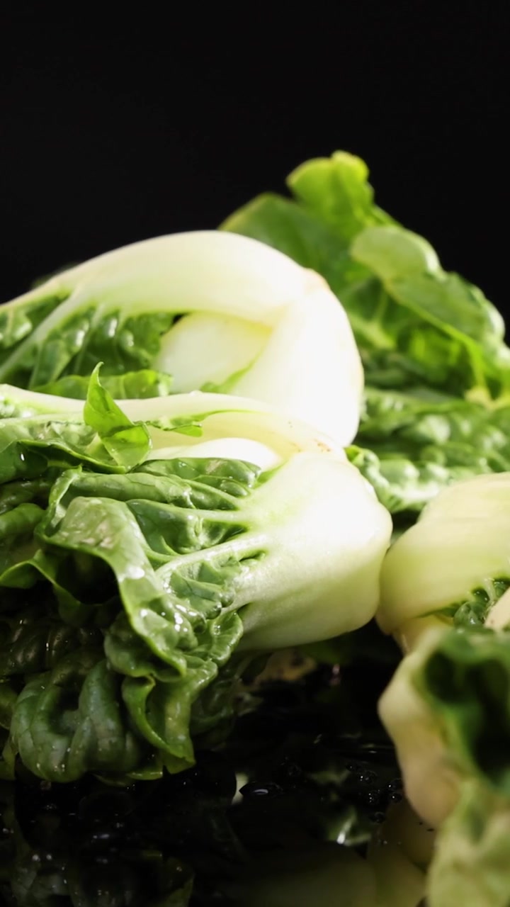 Crisp bok choy falls onto a reflective wet surface, illuminated by dramatic studio lighting against a black background, captured in high-resolution slow motion