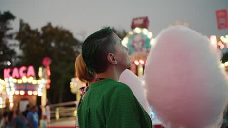 Ein-Mädchen-Mit-Kurzen-Grünen-Haaren-In-Einem-Grünen-Hemd-Und-Ein-Blondes-Mädchen-Mit-Kurzem-Haarschnitt-Halten-Riesige-Rosa-Zuckerwatte-In-Den-Händen-Und-Schauen-Sich-Während-Ihres-Dates-Die-Faszinierend-Leuchtenden-Attraktionen-Im-Vergnügungspark-An