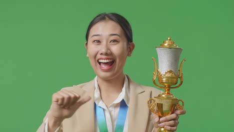 close up of asian business woman in a suit with a gold medal showing gold trophy to camera, smiling, and pointing herself being proud winning as the first winner on green screen background in the studio