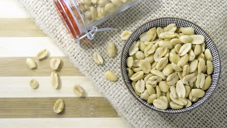 Glass-jar-ceramic-bowl-filled-with-many-peanuts-set-on-woven-hessian-cloth-striped-wooden-table