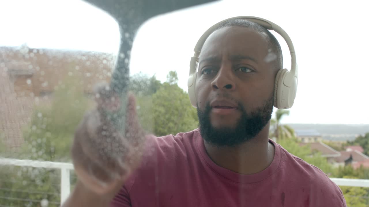 Adult man on balcony wearing headphones, squeezing blue spray bottle, using squeegee to clean glass