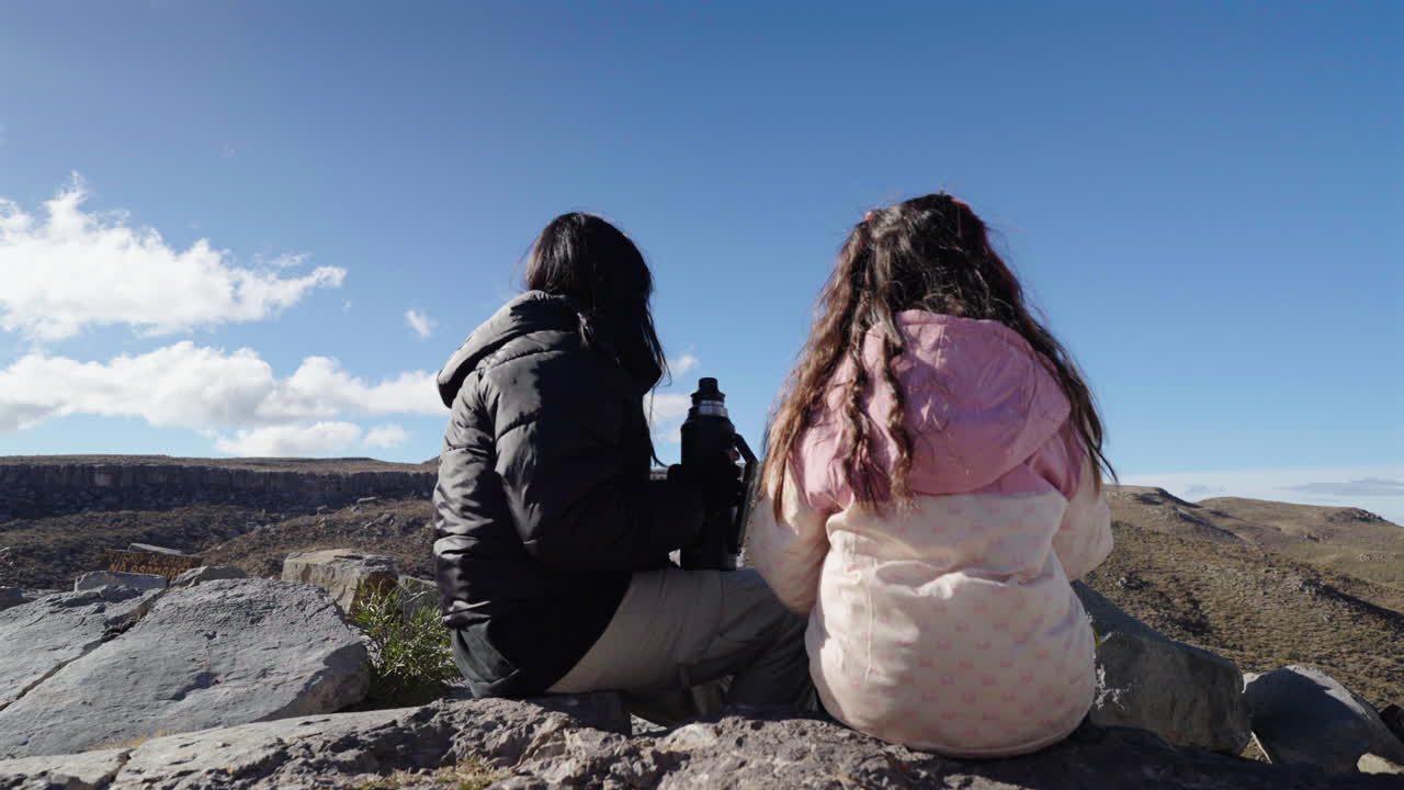 Static shot of a mother and daughter preparing Mate Argentino with Yerba and vacuum flask, outdoors in Malargüe, Mendoza, Argentina, capturing a warm cultural ritual in a highland setting