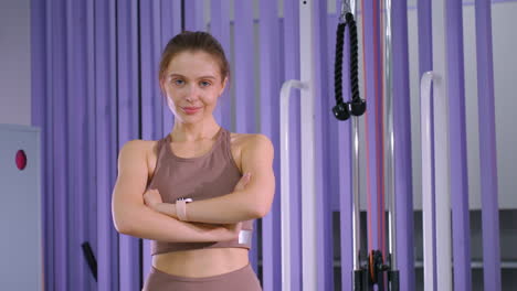 young woman with brown signet hair tied back smiles confidently at camera while standing in gym, she wears athletic wear with folded hands, surrounded by workout equipment