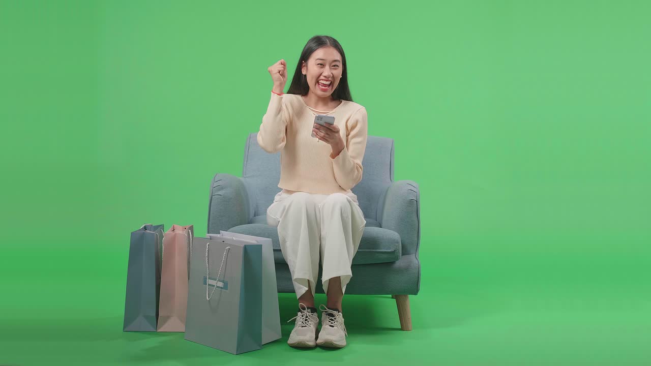 Front View Of An Asian Shopping Woman With Shopping Bags Being Happy Be Able To Using Mobile Phone Shopping Online While Sitting On Sofa In Front Of Green Screen