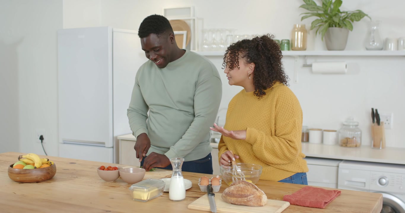 Coppia afroamericana che inizia la preparazione dei pasti, tagliando i pomodori e sbattendo le uova su un'isola da cucina in legno.