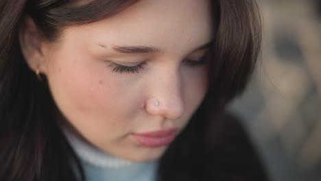 close-up of chic white woman with pink lips, focusing on something in front of her, featuring a subtle nose ring and a golden hoop earring, eyes lowered, soft expression