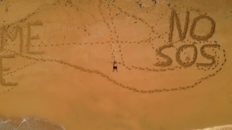 aerial view of a man lying on a desert beach with inscriptions in the sand saying leave me here and no sos, in são tomé,africa - rising drone shot