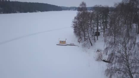 Luftaufnahmen,-Die-Eine-Weite,-Weite-Winterlandschaft-Zeigen,-Während-Gleichzeitig-Sanft-Auf-Eine-Kleine-Hütte-Am-Waldrand-Gezoomt-Wird