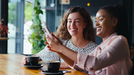 Two-Young-Female-Friends-Meeting-In-Coffee-Shop-And-Posing-For-Selfie