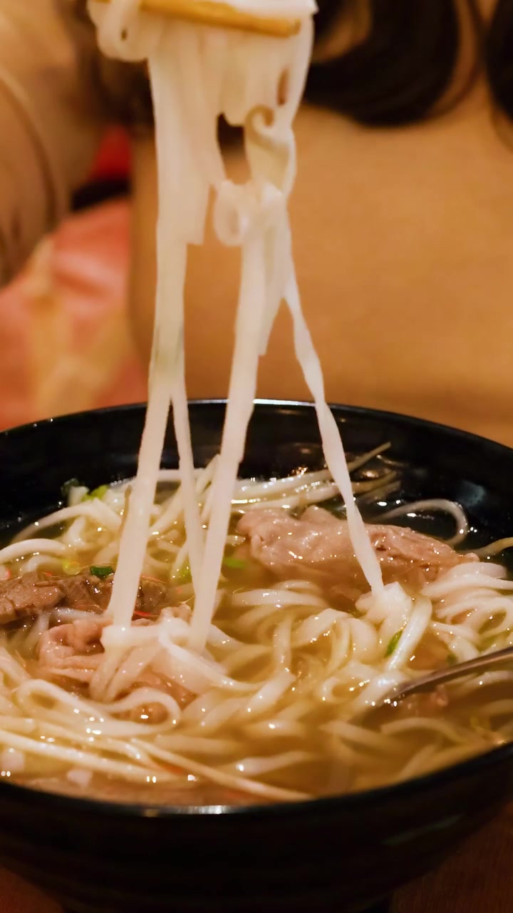 Una persona solleva abilmente i noodles da una ciotola fumante di pho vietnamita usando le bacchette in un ambiente illuminato calorosamente