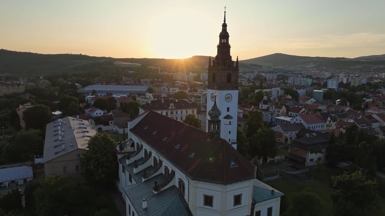 Wide drone zoom out showing the church tower and full Litomerice city under a colorful sky at sunset.