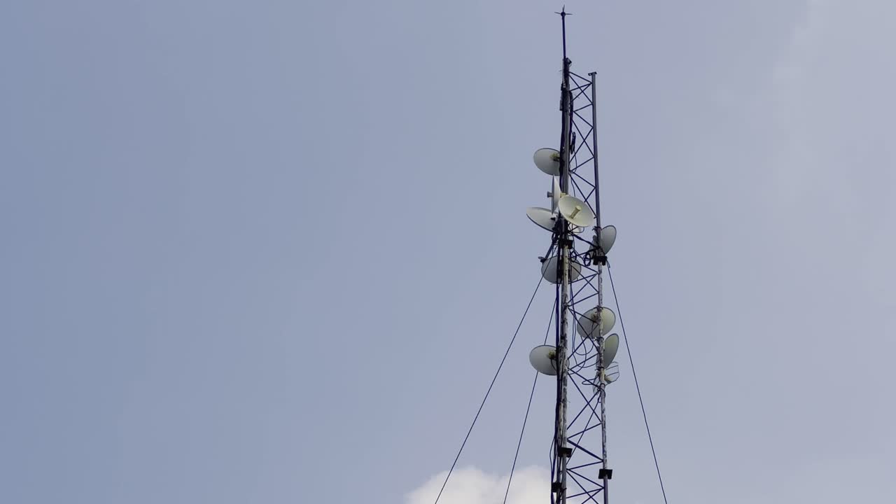 camera tilts up to showcase a towering metal structure, featuring numerous satellite dishes, set against blue sky