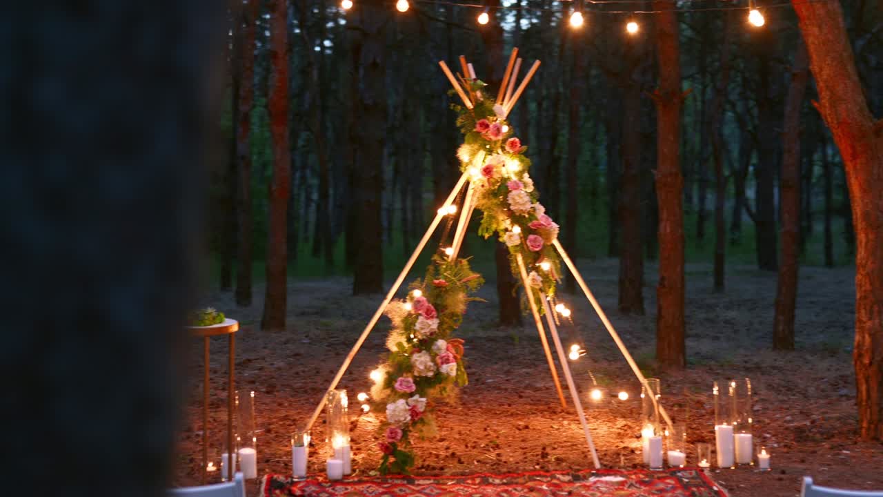 Bohemian tipi wooden arch decorated with burning candles, roses and pampass grass, wrapped in fairy lights illumination on outdoor wedding ceremony venue in pine forest at night. Bulbs garland shines
