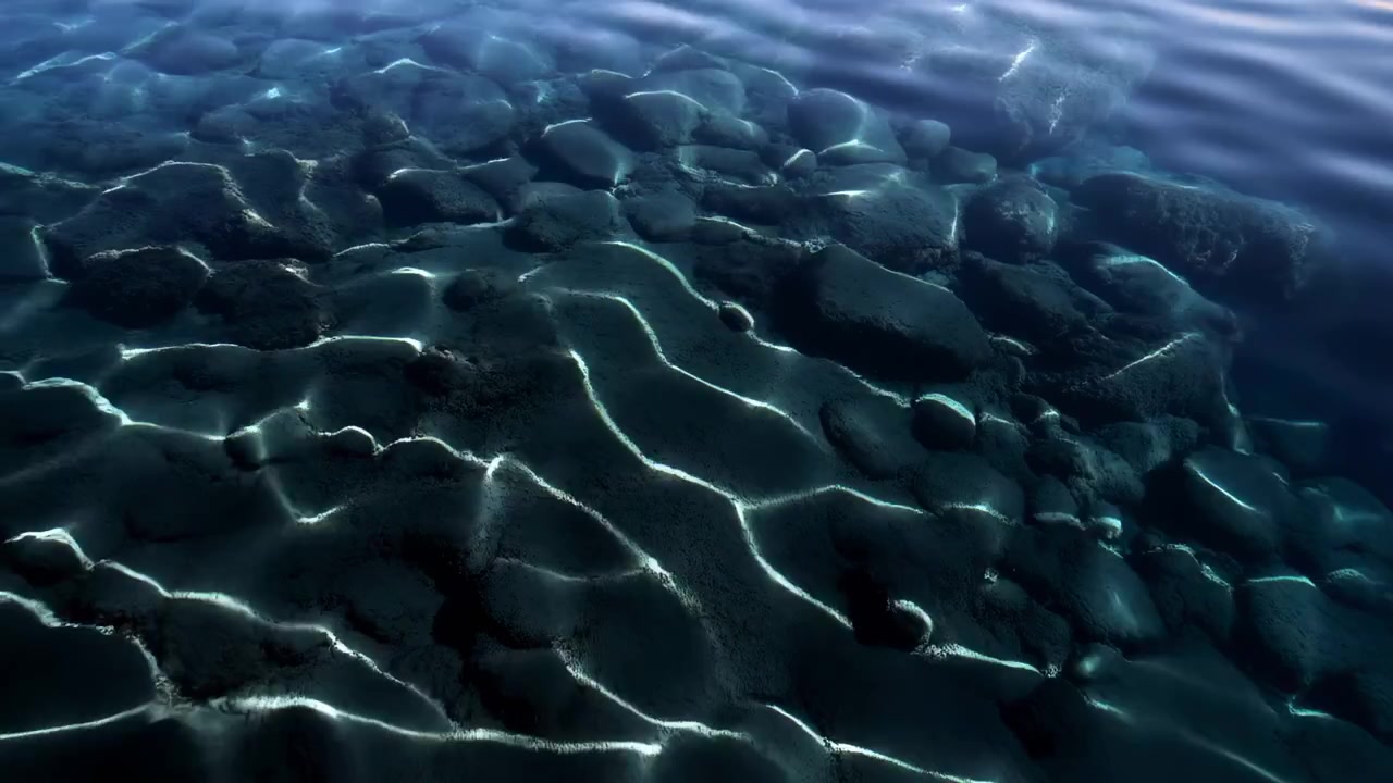 Wide oblique aerial motion scene showing shallow underwater seabed composed of rounded boulders and smooth ridges visible through calm, clear blue-green water