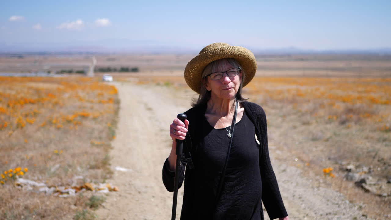 A happy middle aged woman with a hat and walking stick hiking down a dirt road with orang flowers in slow motion
