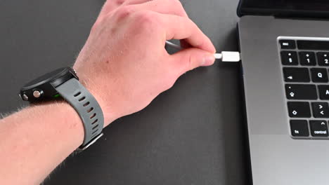 an electronic device user plugs a cable into his computer to charge it with a watch at its wrist, use of a laptop with a keyboard