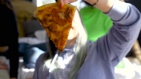 close up of a girl holding a slice of pizza and eating it while her friend recording a video