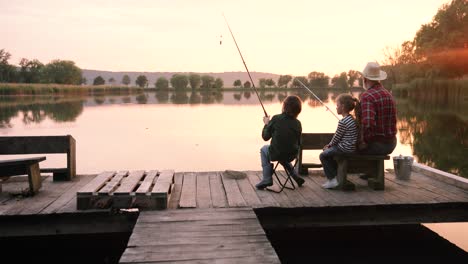 cámara que se acerca desde la parte trasera de un niño y una niña adolescentes sentados con su abuelo en el muelle del lago y pescando juntos