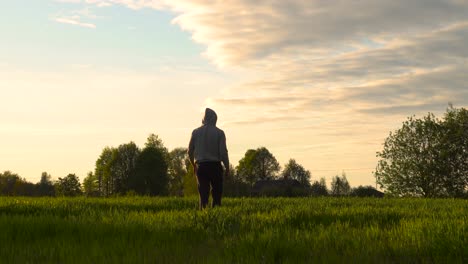 Männlicher-Spaziergang-Auf-Grünem-Grasland-Während-Des-Malerischen-Sonnenuntergangs-Zur-Goldenen-Stunde-Mit-Wolken