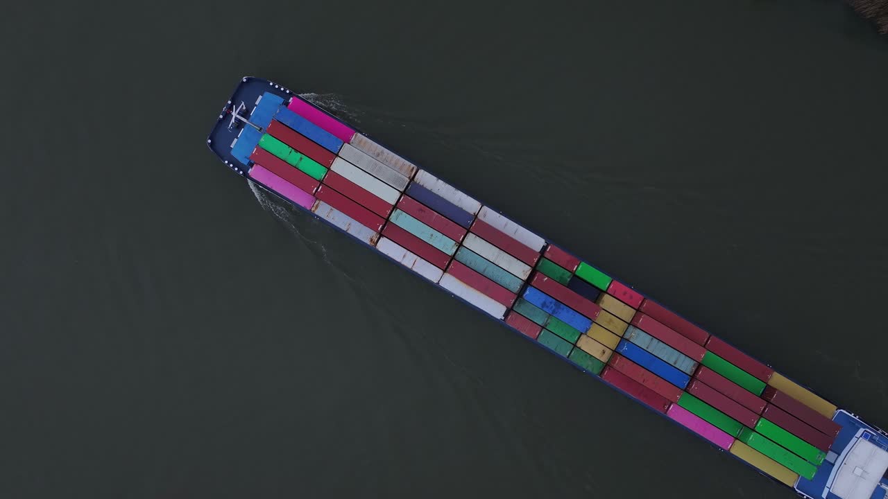 Cargo ship with red, green, blue and yellow containers cuts a dark ribbon through the Puttershoek channel under soft evening light with its geometric pattern, top down overhead drone shot