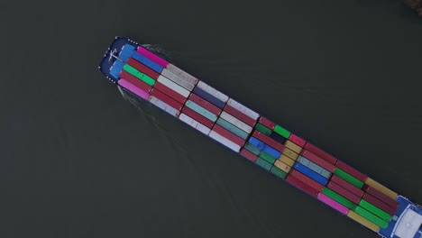 cargo ship with red, green, blue and yellow containers cuts a dark ribbon through the puttershoek channel under soft evening light with its geometric pattern, top down overhead drone shot
