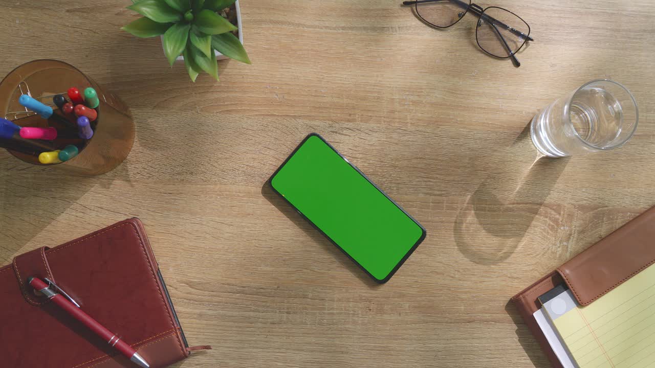 vista de arriba hacia abajo de una computadora portátil con pantalla verde simulada pantalla chromakey en un escritorio de oficina de madera al lado de la libreta con bolígrafos, gafas y un vaso de agua.