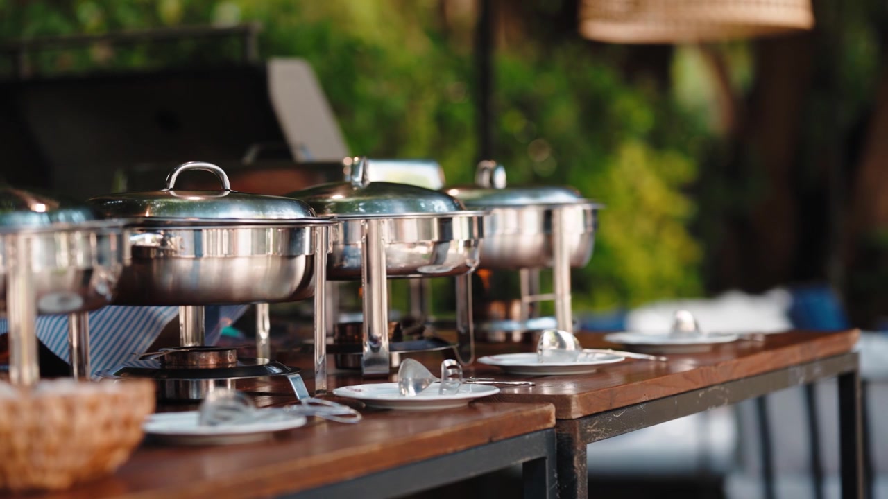 A row of chafing dishes with lids and a bread basket on a wooden buffet table outdoors, with plates set for guests.