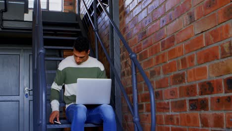 Mann-Spricht-Auf-Dem-Handy,-Während-Er-Auf-Der-Treppe-Einen-Laptop-Benutzt,-4K