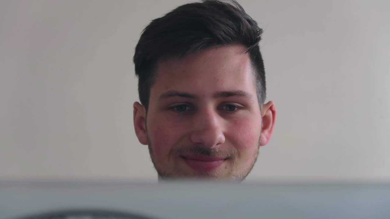 un joven trabajando en una computadora y mirando la pantalla, pensando y sonriendo, con una computadora portátil en primer plano, todavía filmado en primer plano en ángulo bajo