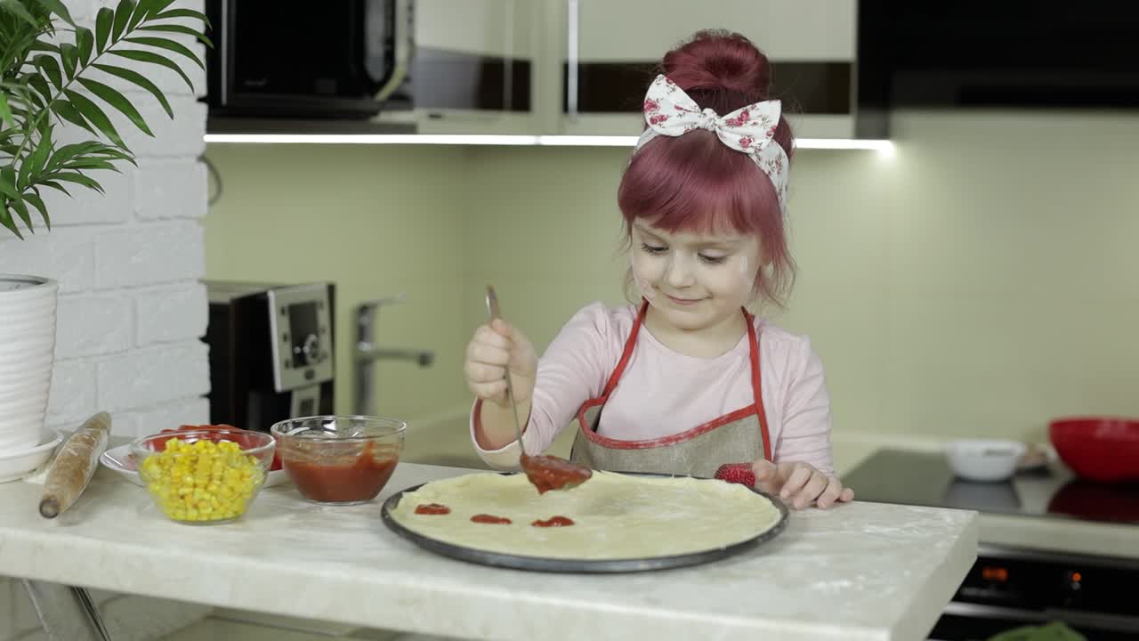 cocinando pizza. niño pequeño en delantal añadiendo salsa de tomate a la masa en la cocina