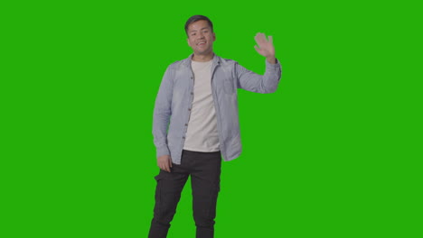 Portrait-Of-Casually-Dressed-Smiling-Young-Man-Waving-In-Welcome-Against-Green-Screen-