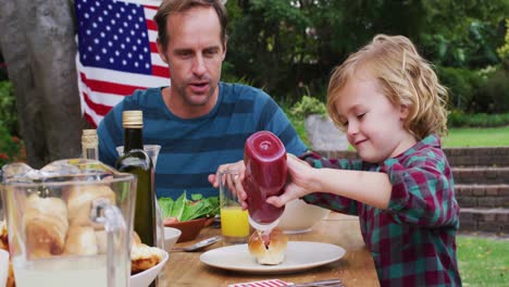 Smiling-caucasian-man-watching-son-using-ketchup-during-celebration-meal-in-garden