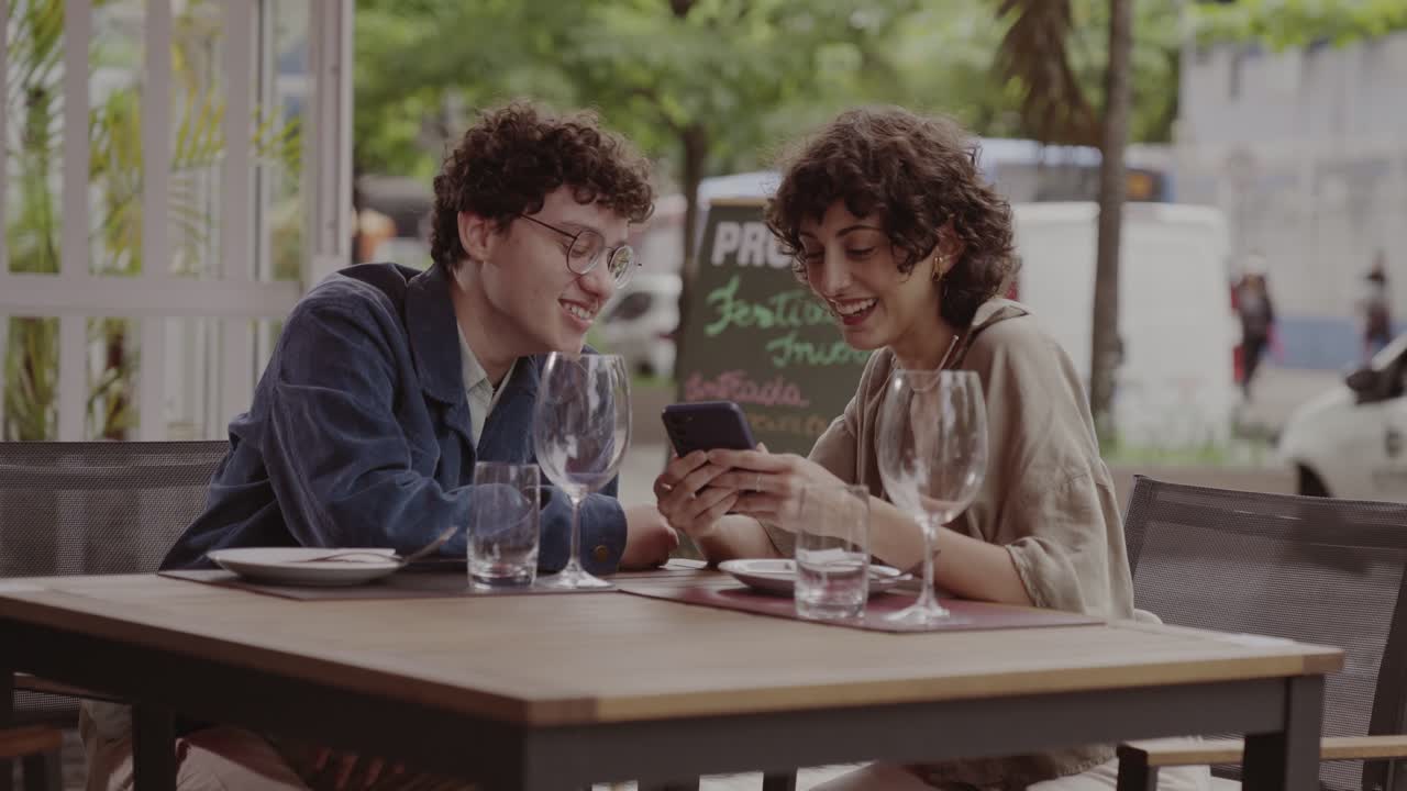 Two friends or a couple sharing a moment and a smartphone at an outdoor cafe