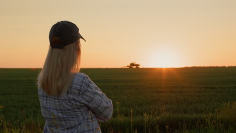 a successful woman farmer in the city looks at her wheat field, where a tractor is working in the distance. silhouette at sunset