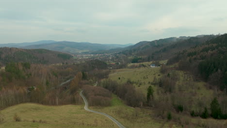 a winding road cuts through a lush valley surrounded by hills and sparse forest, with a village in the distance under a cloudy sky